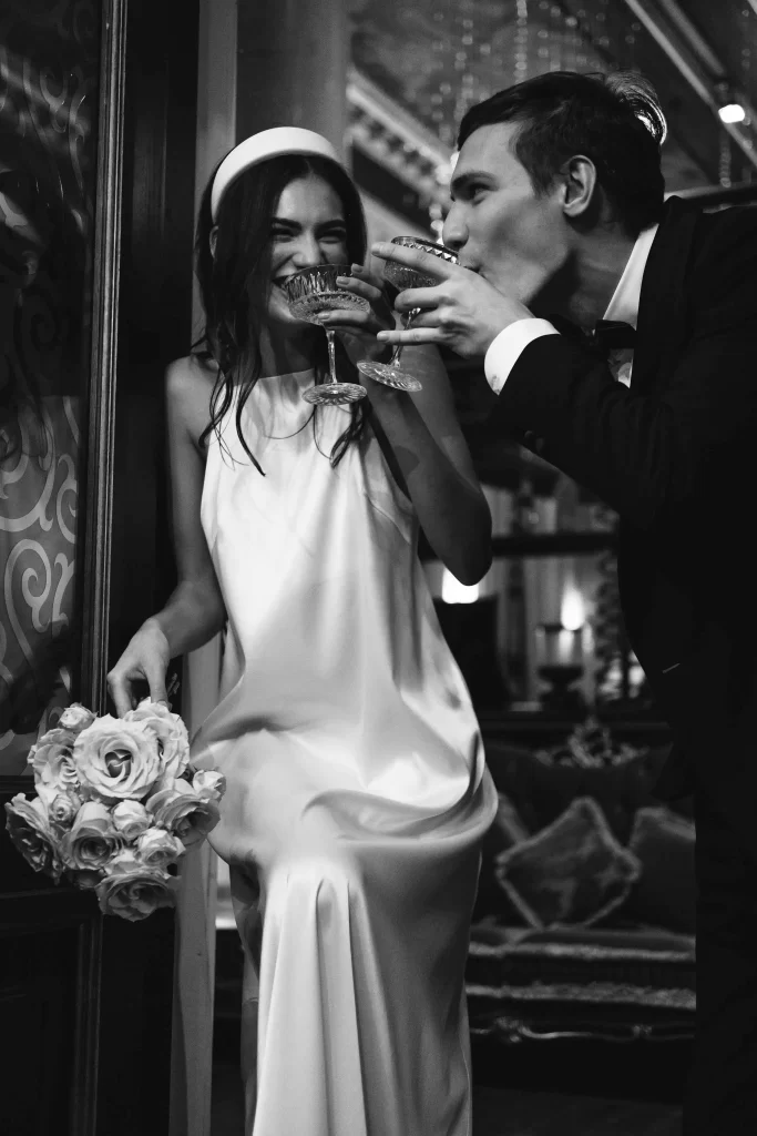 wedding couple drinking,Elegant wedding dress with intricate lace details hanging in a rustic stone room with a wooden beam ceiling and soft natural lighting